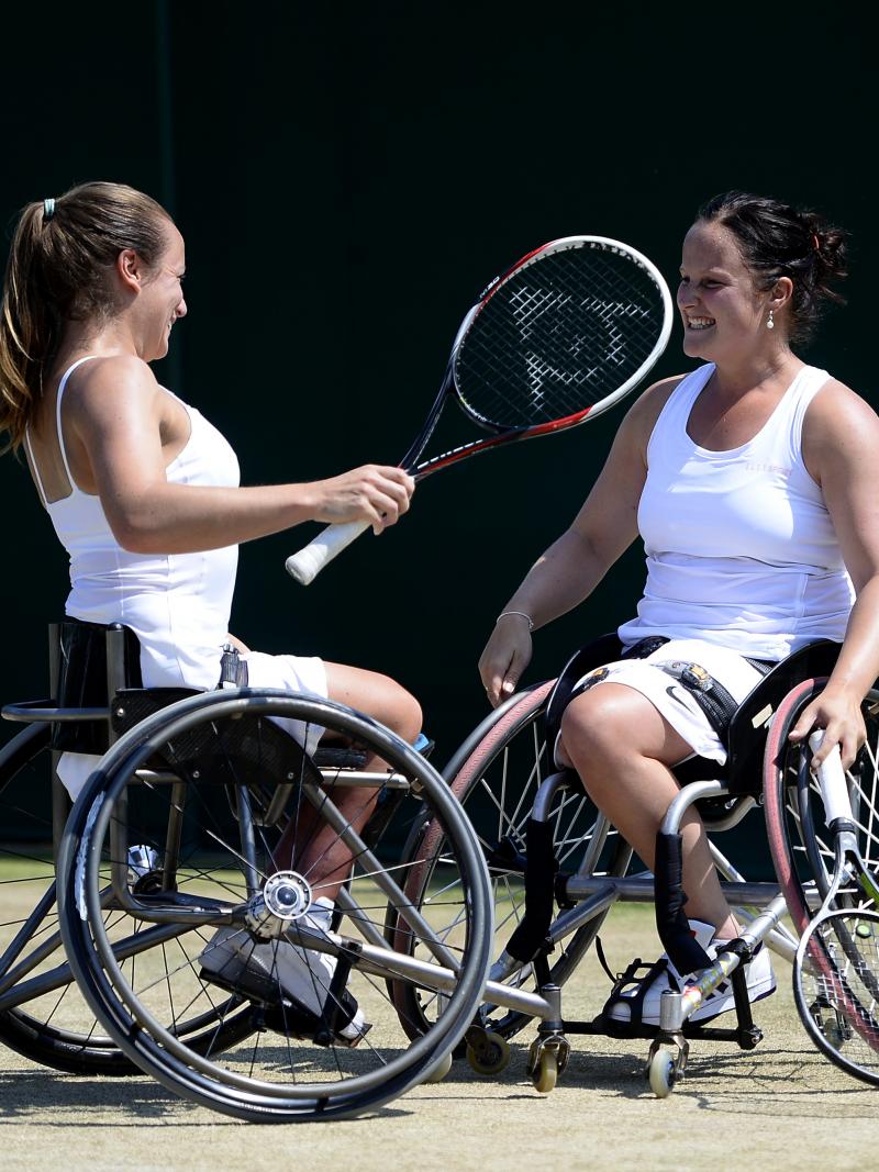 Two women in wheelchairs look at each other smiling on a grass tennis court.