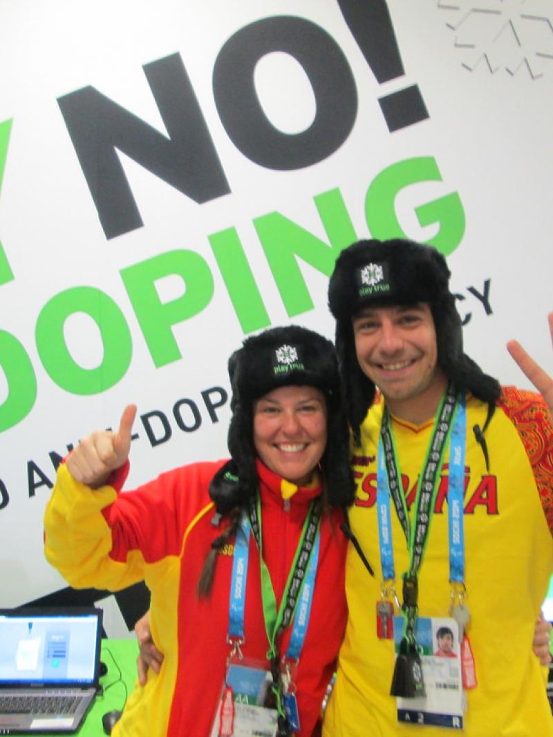 Two people give a thumbs up and a peace sign in front of a "Say No! to Doping" sign.
