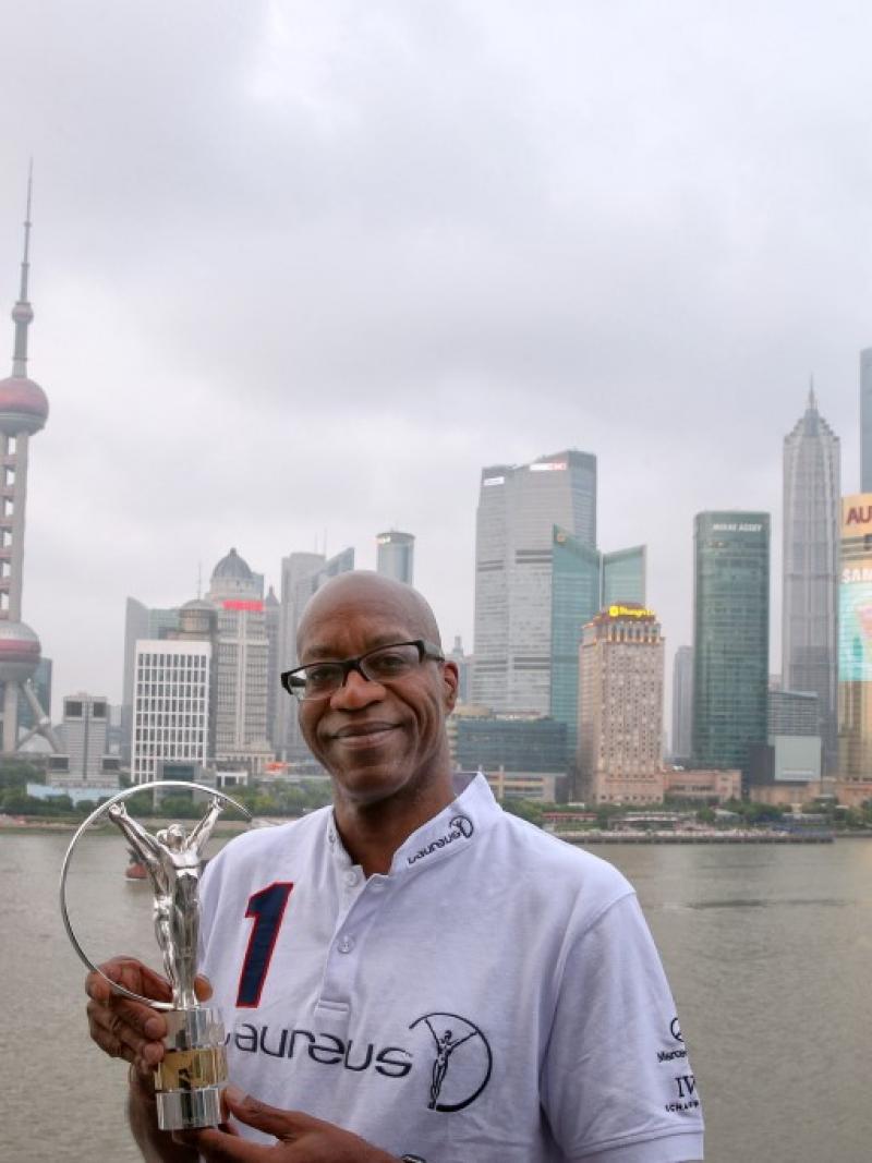 A man's upper body in front of a skyline with skyscrapers in the background. He is holding a silver statue to the camera, smiling