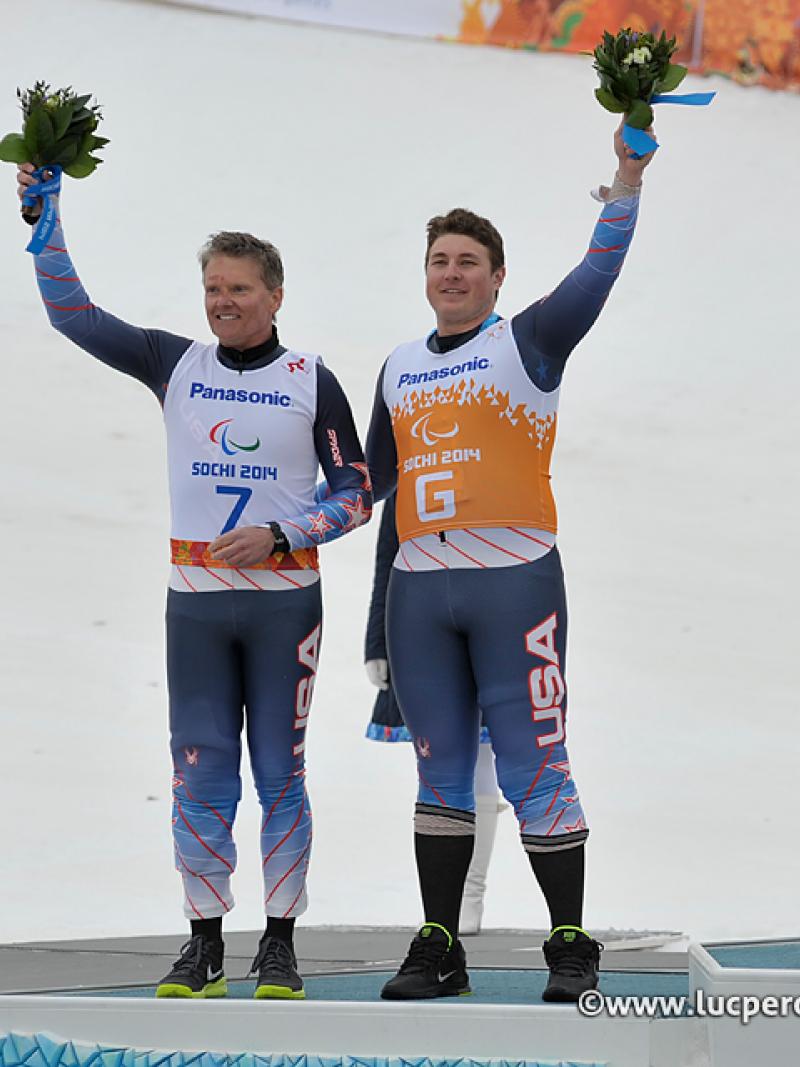 Two men in skiing suits waving flower bouquets. They both stand on a podium in the snow