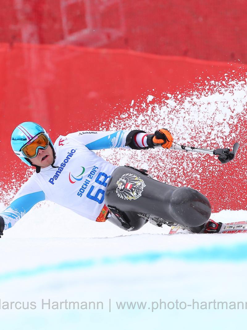 Sit skier Roman Rabl rounds a turn very low, at speed with snow spraying behind him and outriggers outstretched.