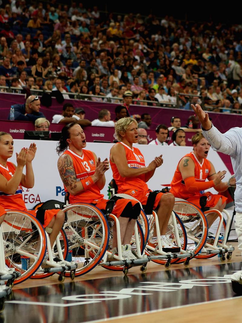 Female wheelchair basketball players lined up, cheering during a game with an amputee man in a suit standing in front of them