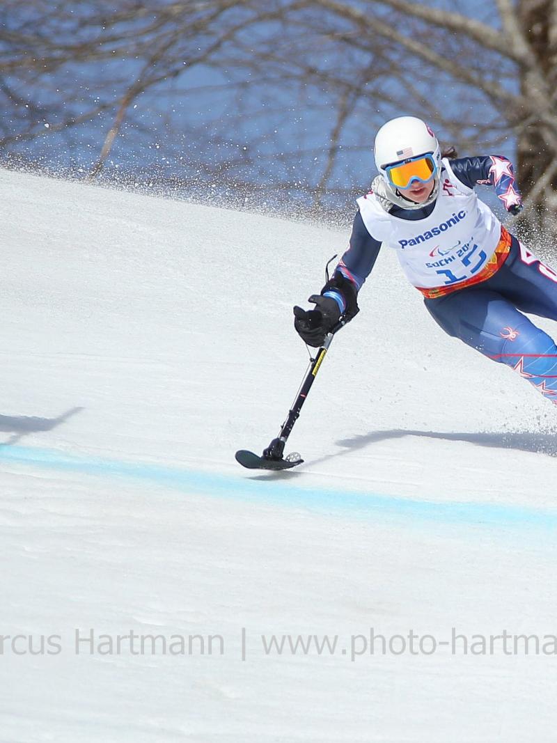 Skier with one leg skis at speed past a gate with her outrigger outstretched