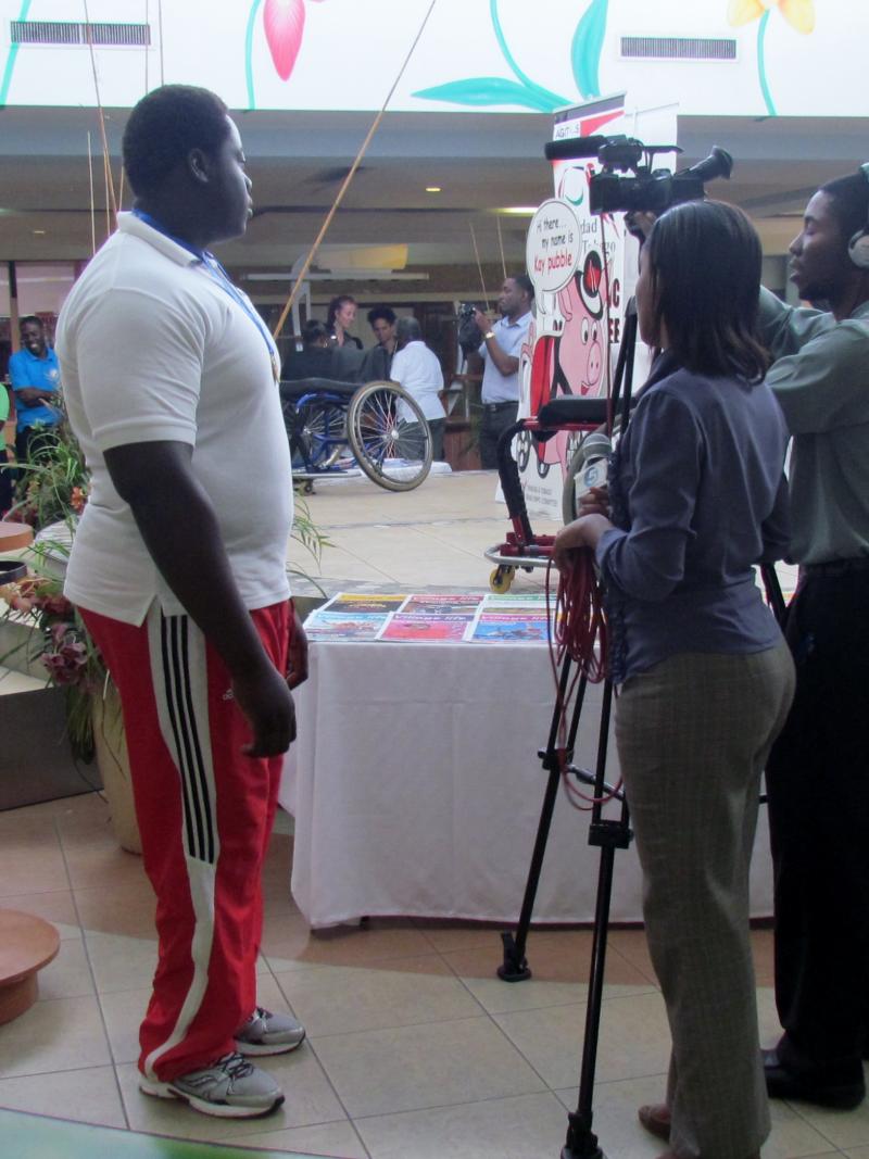 Team of a camera man and a women holding a microphone stand opposite of a man in sport dress. They seem to be preparing an interview. Background: A stage with a wheelchair on it