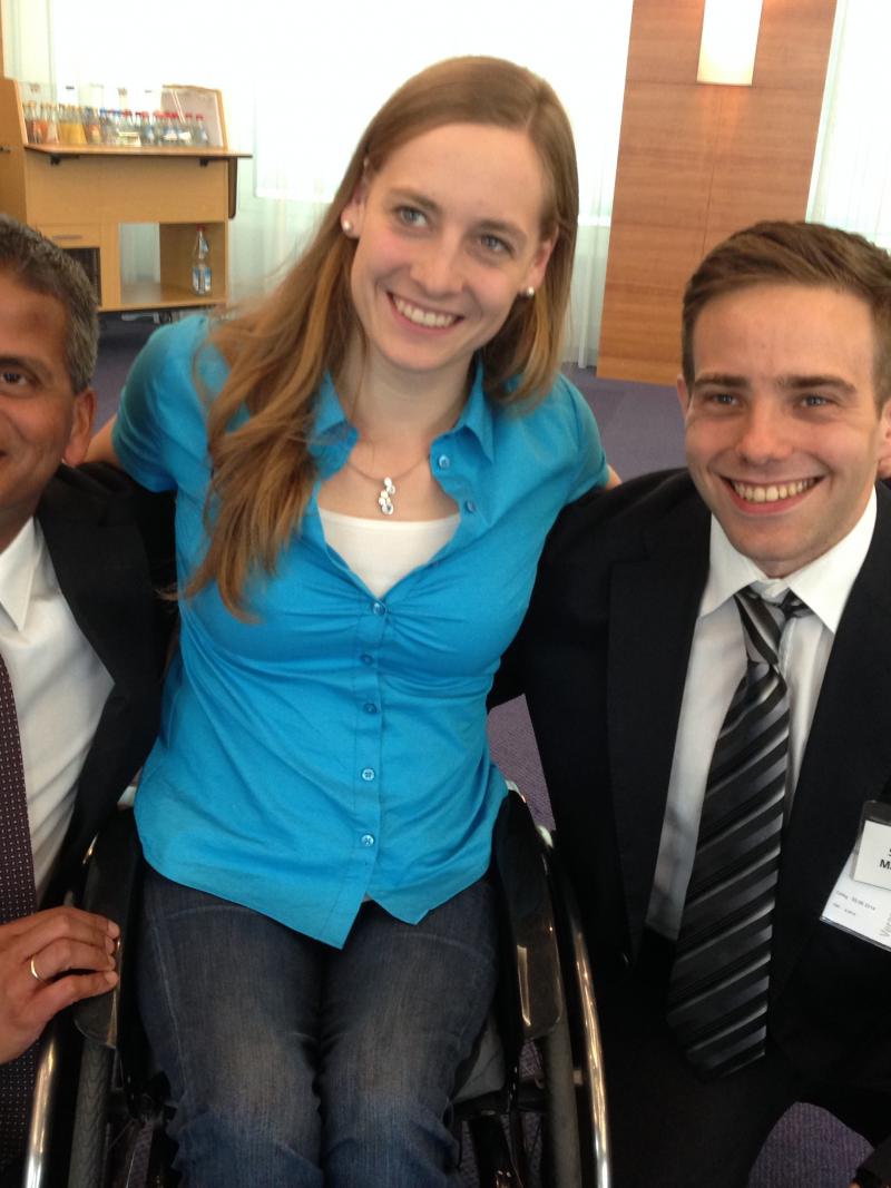 Women in wheelchair smiling, poses surrounded by two men in suits who perch next to he