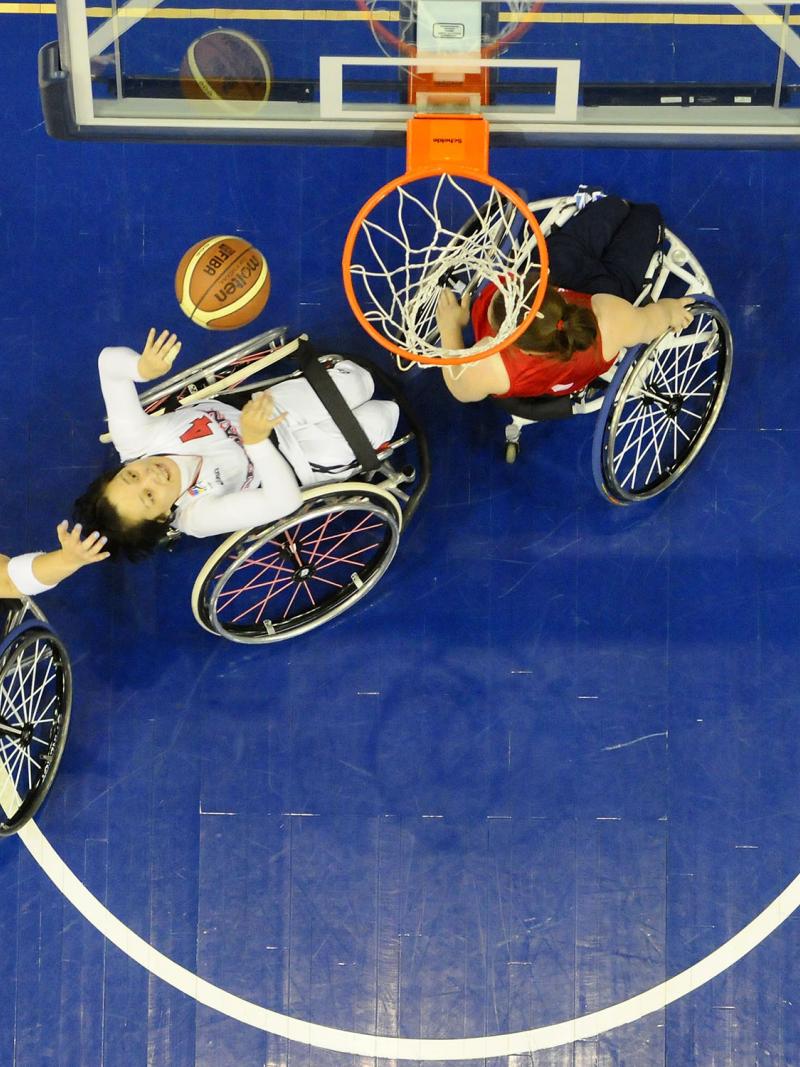 Picture taken from above the basketball basket. Three female players in wheelchairs fighting for the ball under the basket.