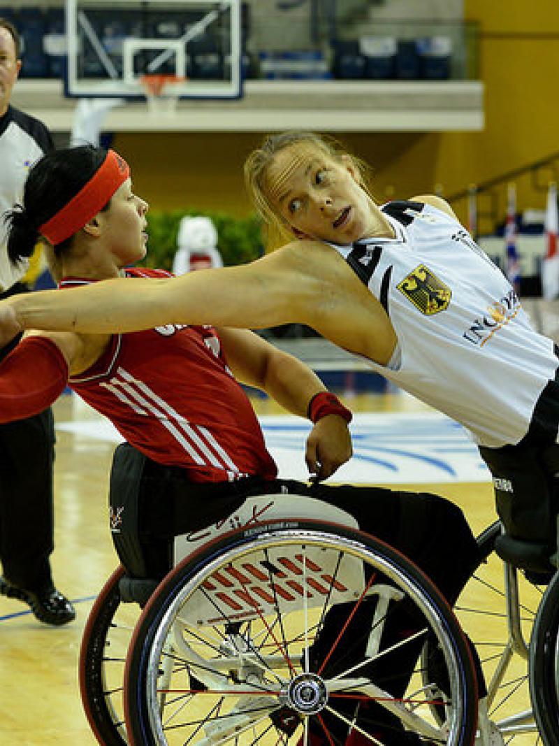 Two female basketball players in wheelchairs fight for the ball. A referee watches them from behind