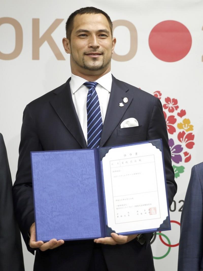 Three men standing next to each other, looking at the camera. Background: Tokyo 2020 logo. Athletic man in the middle showing a document to the camera