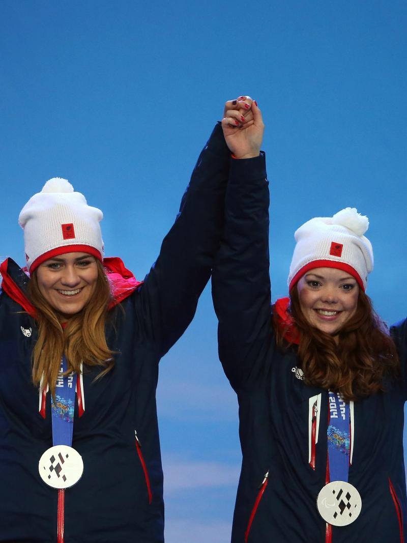 Two young women wearing ski clothes and a silver medal holding hands and waving