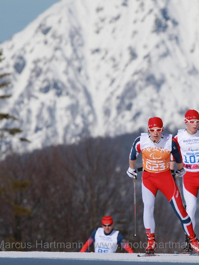 Wide shot of two Nordic skiers skating close together on the crest of the hill, with one skier a short distance behind. Big mountain in background