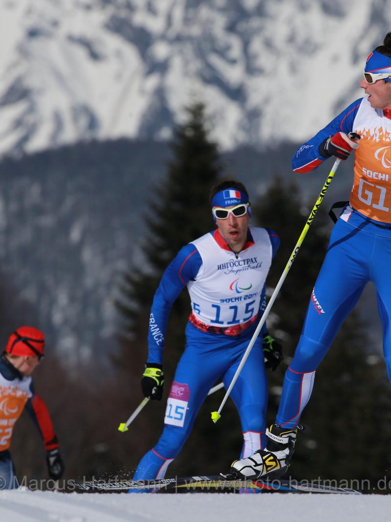 A Nordic skier looks behind him at two other skiers approaching the crest of the hill, close behind him. Mountains are in the background.
