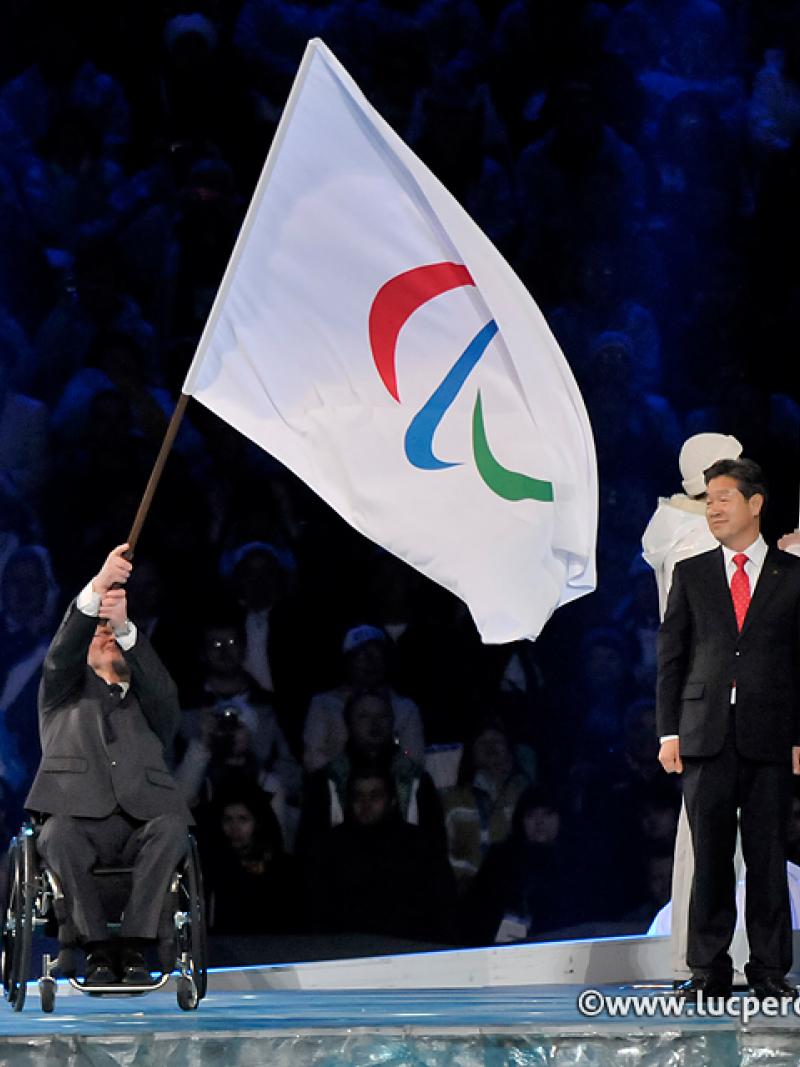 Man in wheelchair on a stage waving a huge flag with the Paralympic symbol on it, handing it over to another person.