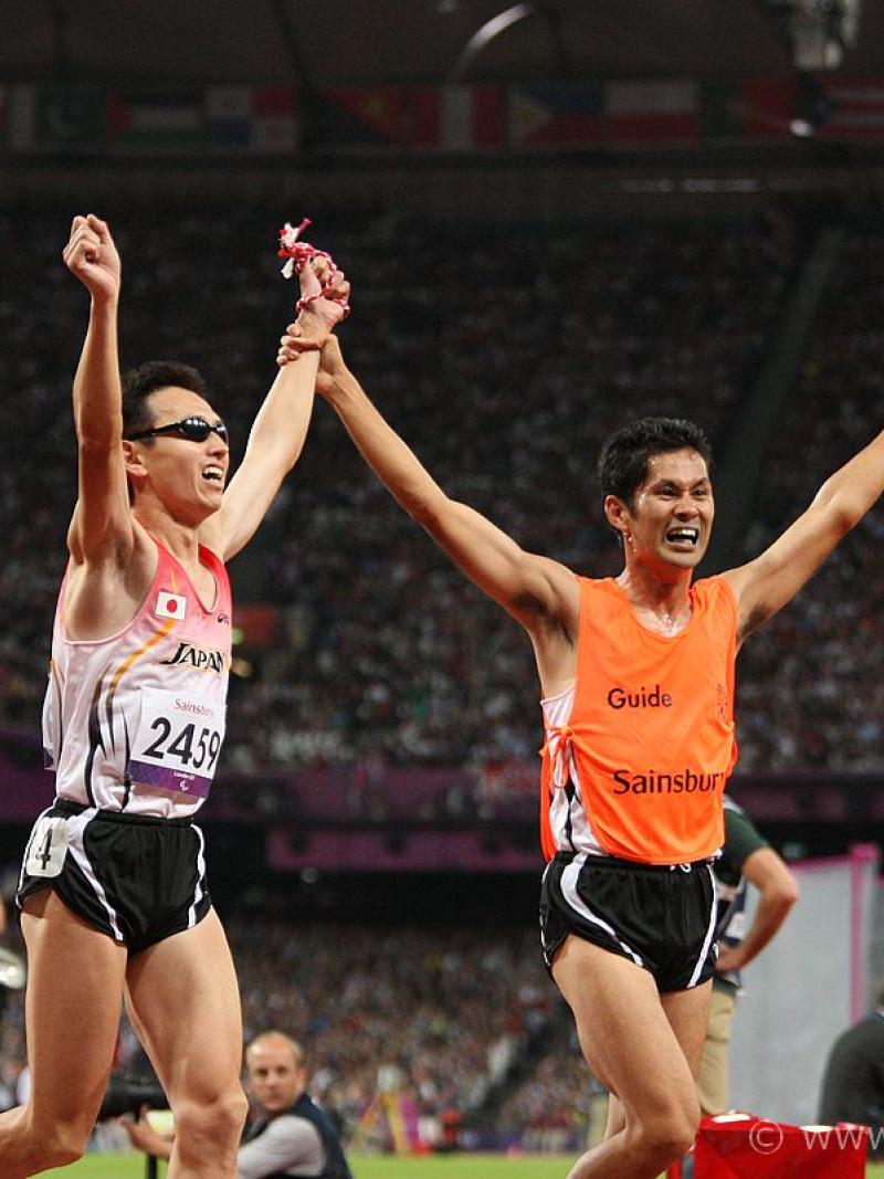 Runner with a guide crossing the finish line with their hands in the air
