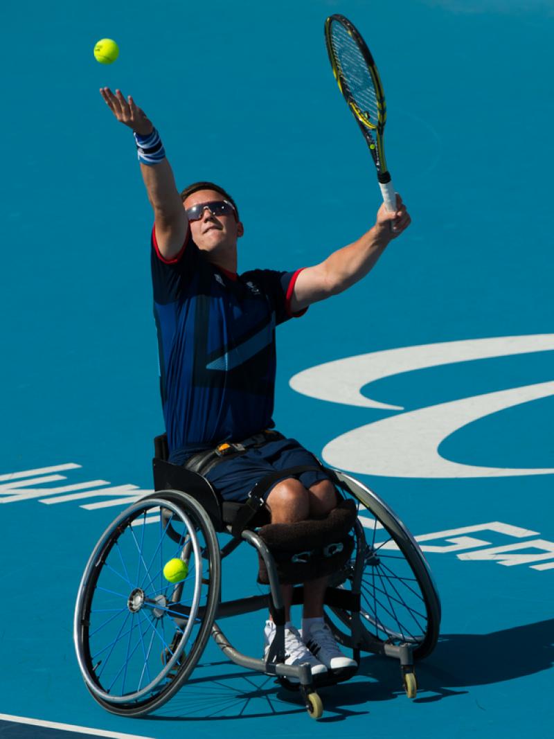 Man in wheelchair serves a ball on a tennis court.