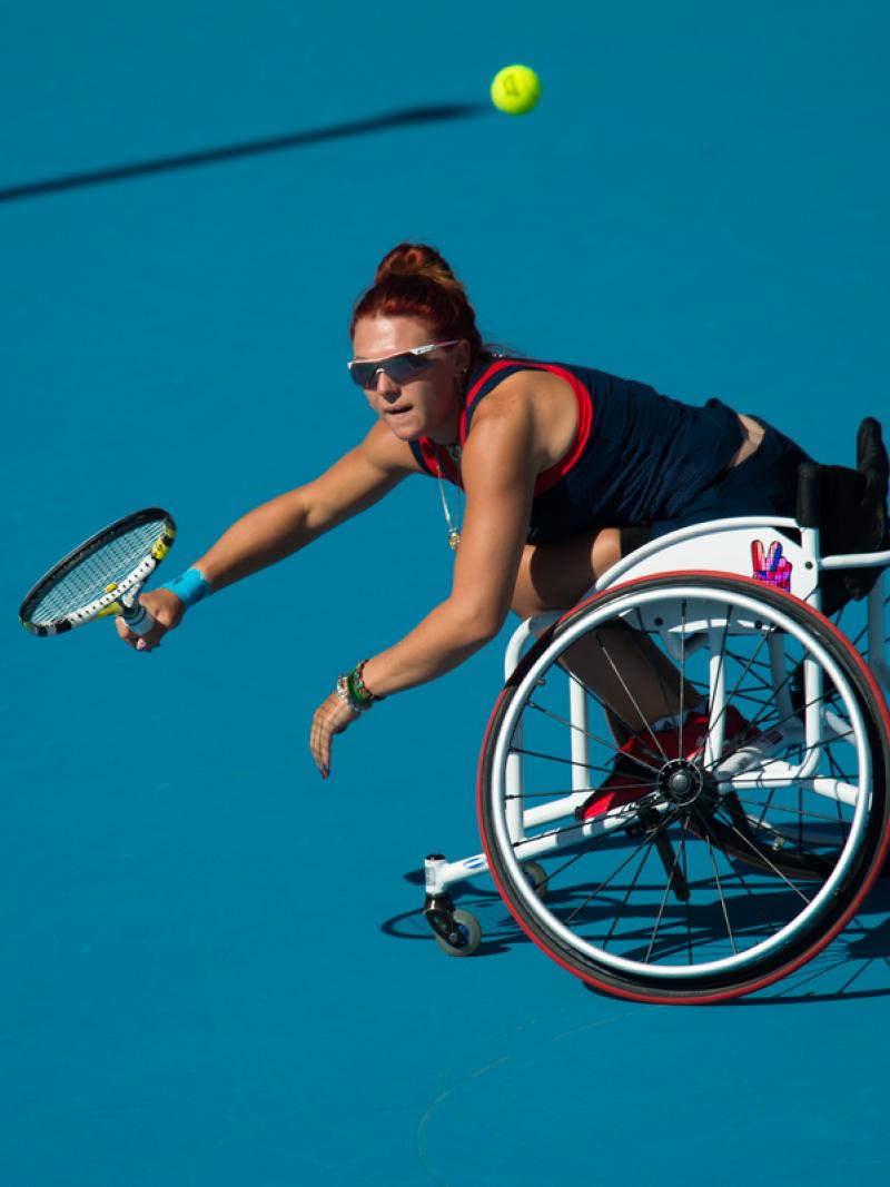 Woman in a wheelchair on a blue tennis court trying to reach a ball