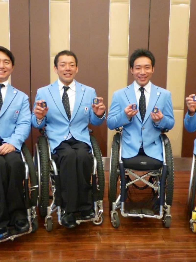 Four men in wheelchairs all wearing blue jackets show pins to the camera, smiling