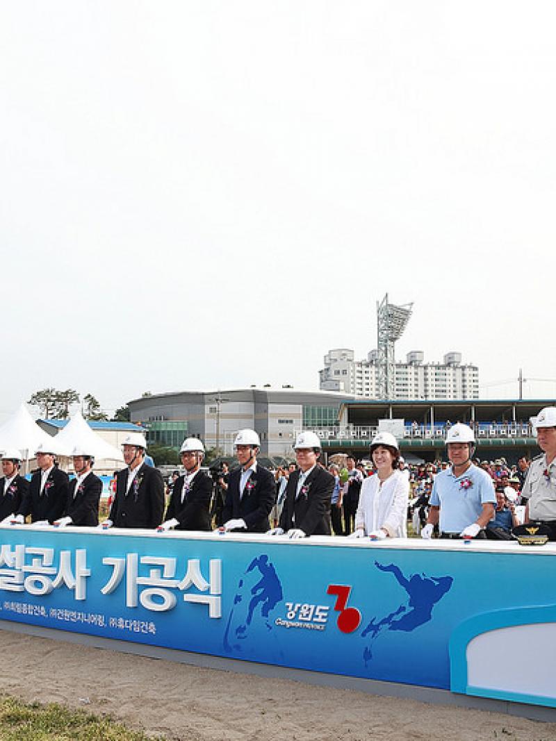 People in suits with construction helmets stand behind a banner in front of a construction area