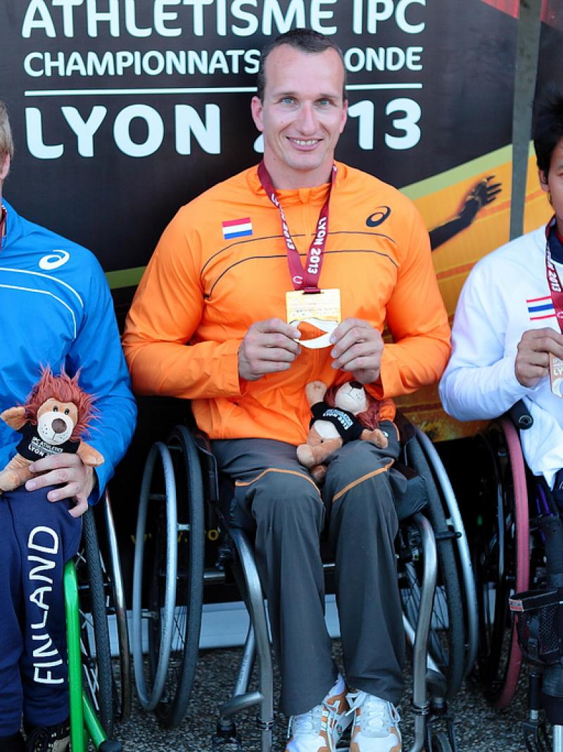 Three wheelchair athletes pose with their medals in front of a Lyon branded background