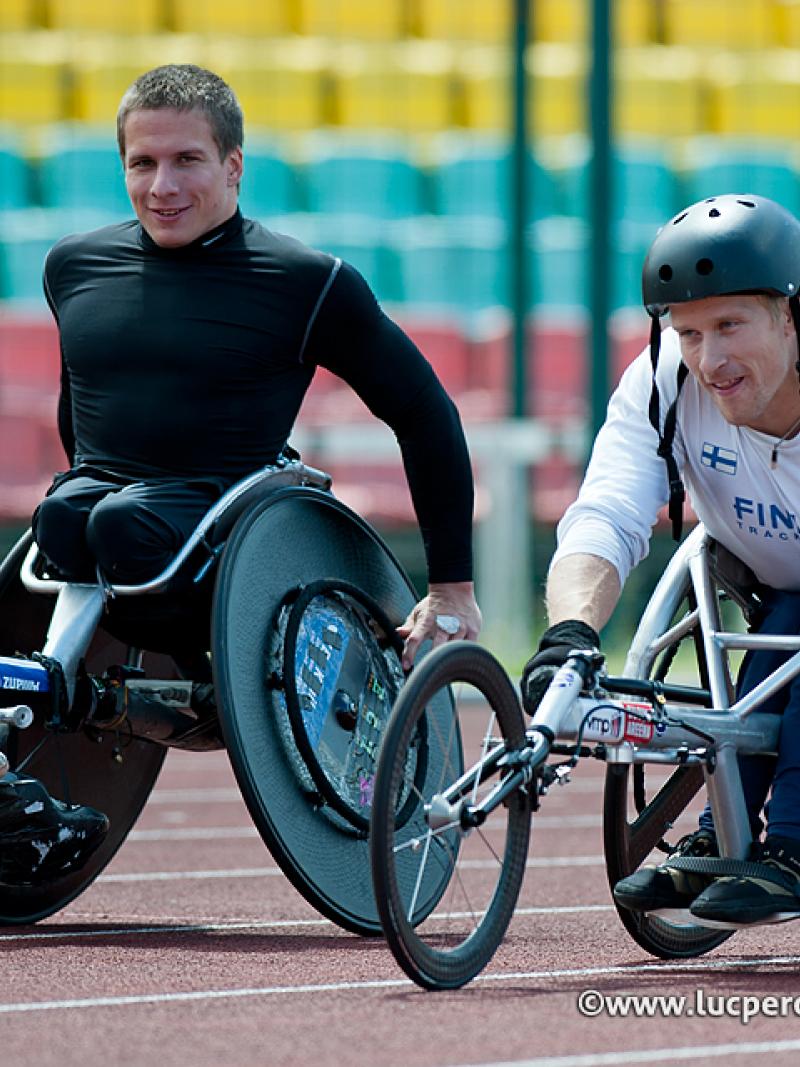 Two male athletes in racing wheelchairs on track, smile to the camera