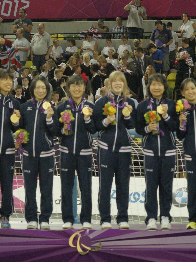 Six women in training suits on podium with medals and flower boquets.