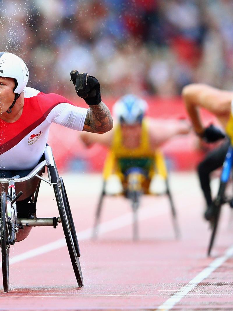 Man in racing wheelchair crosses finish line of a track throwing hands in the air, celebrating