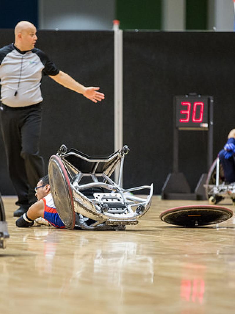 Dressed in green and seated in a wheelchair, a player looks on as another lies face down with his wheel broken from his chair.