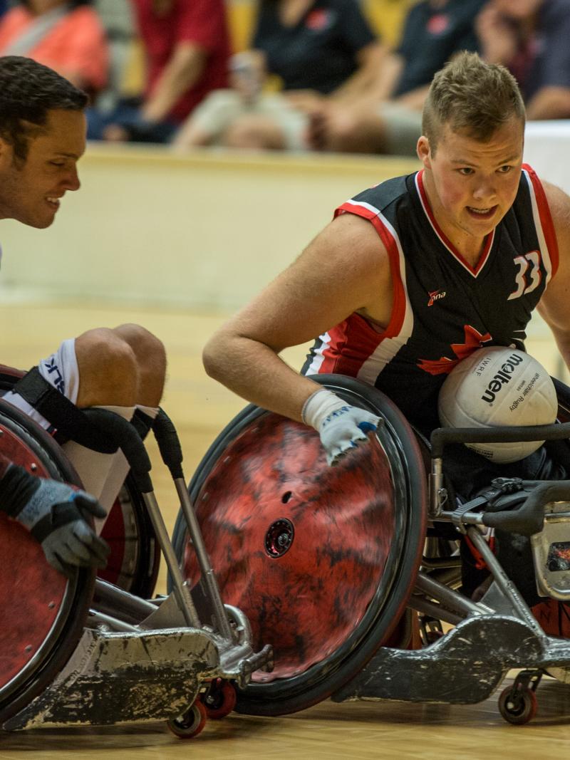 A player wearing blue seated in a wheelchair with a white ball on his lap about to collide with an opponent wearing white.