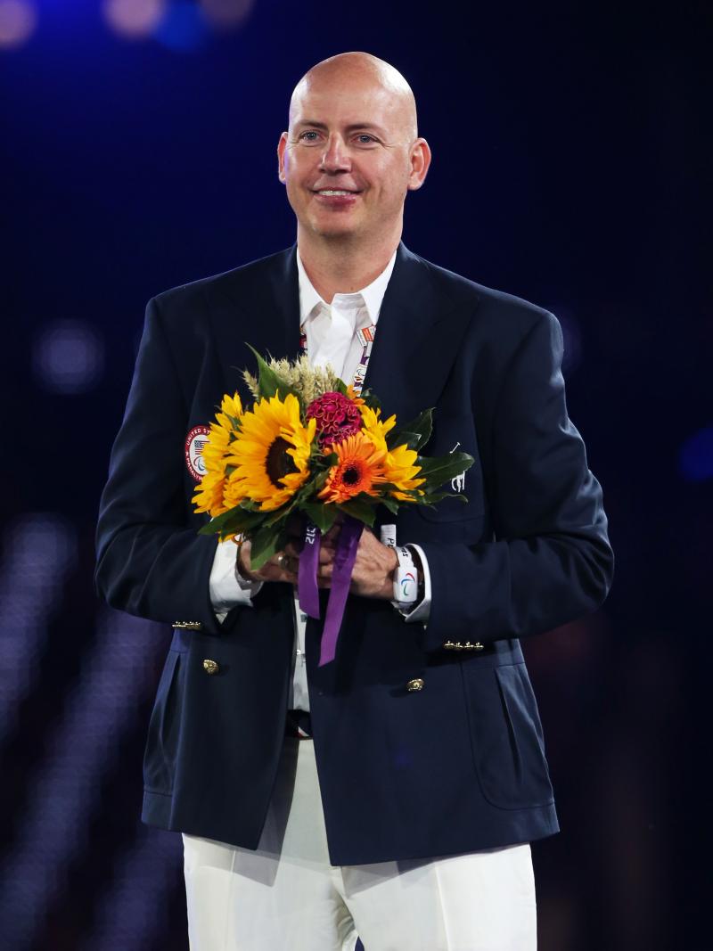 A picture of a smartly dress man, with light trousers and dark jacket holding a bunch of flowers.