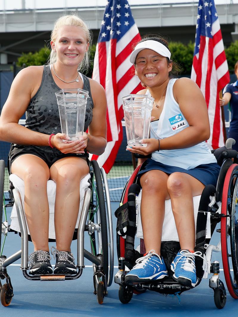 Two players sat in wheelchair holding trophies on their laps. In the background are two USA flags.