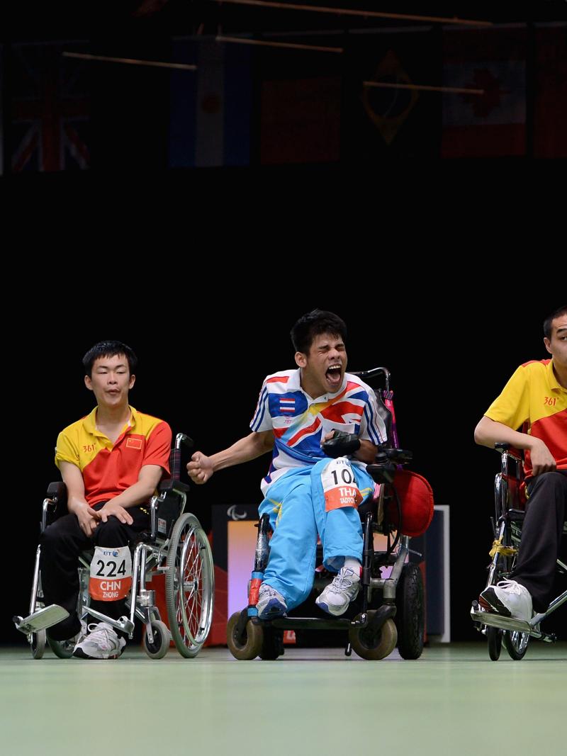Four men in wheelchairs and one man standing on a field of play with dark background. Three men celebrating