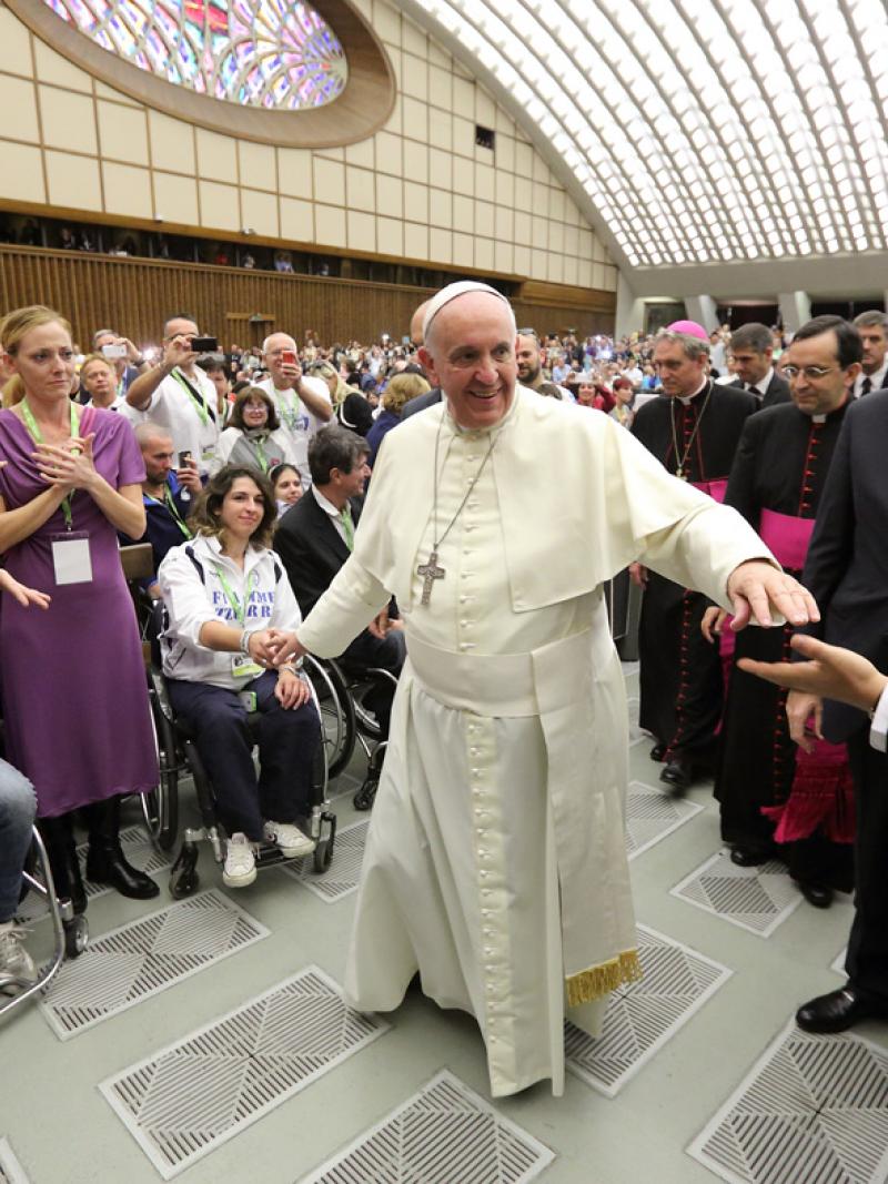 Man in white robe walks through a crowd of people, some of them in wheelchairs