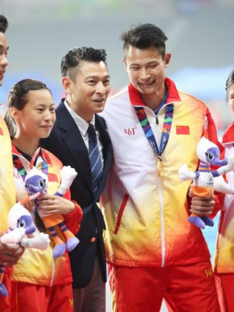 Man in dark suit stands between some athletes on the podium
