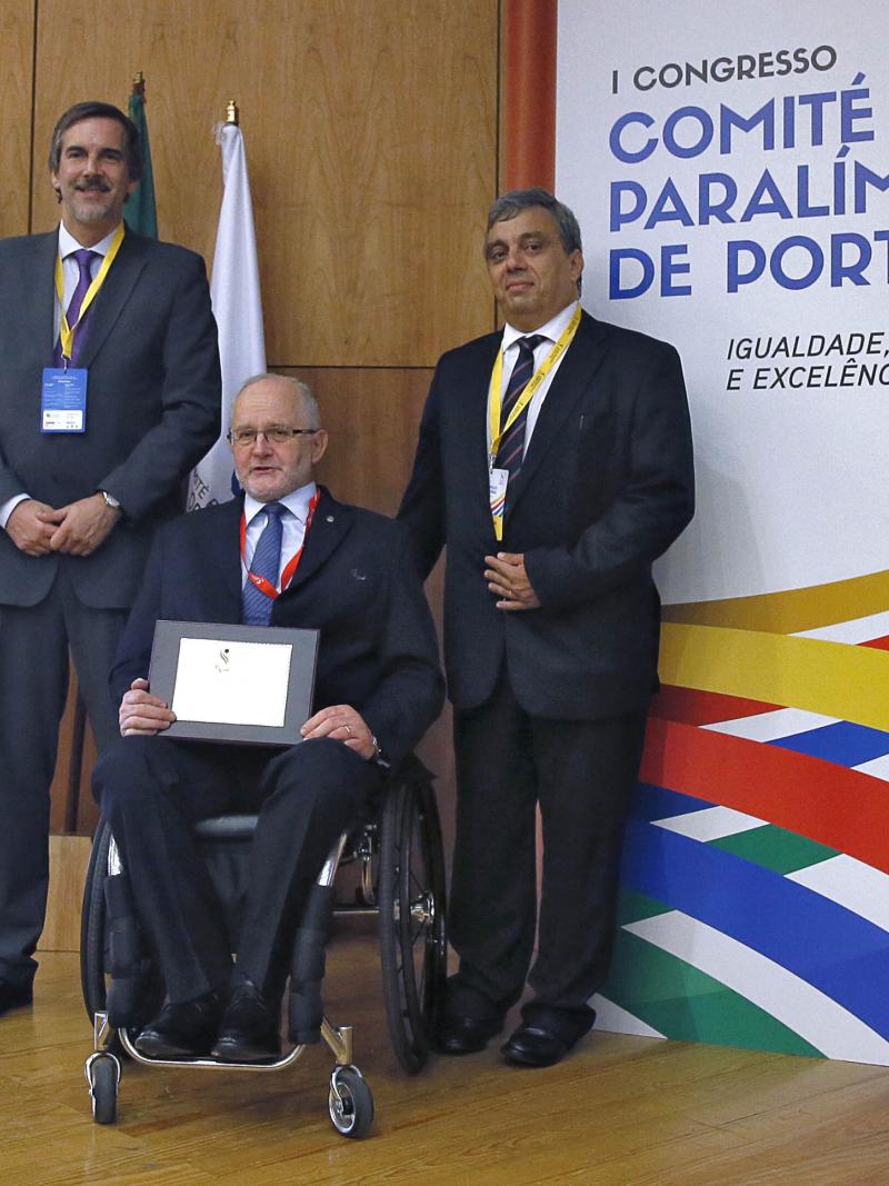 Three men (one in a wheelchair) pose in front of a wooden wall and some flags
