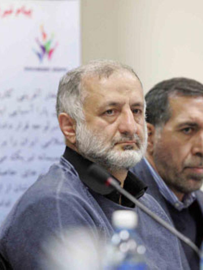 Upper bodies of two men sitting behind a table, listening to something.