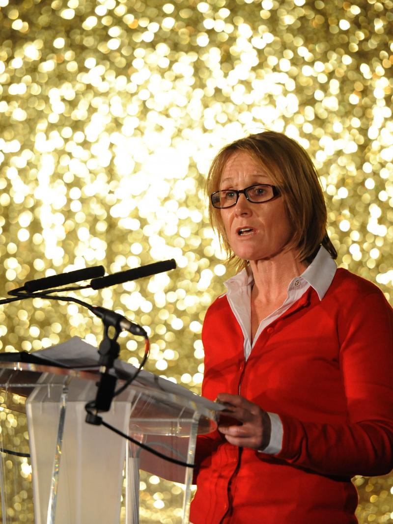 woman in red standing on stage in front of a gold background and addressing the crowd