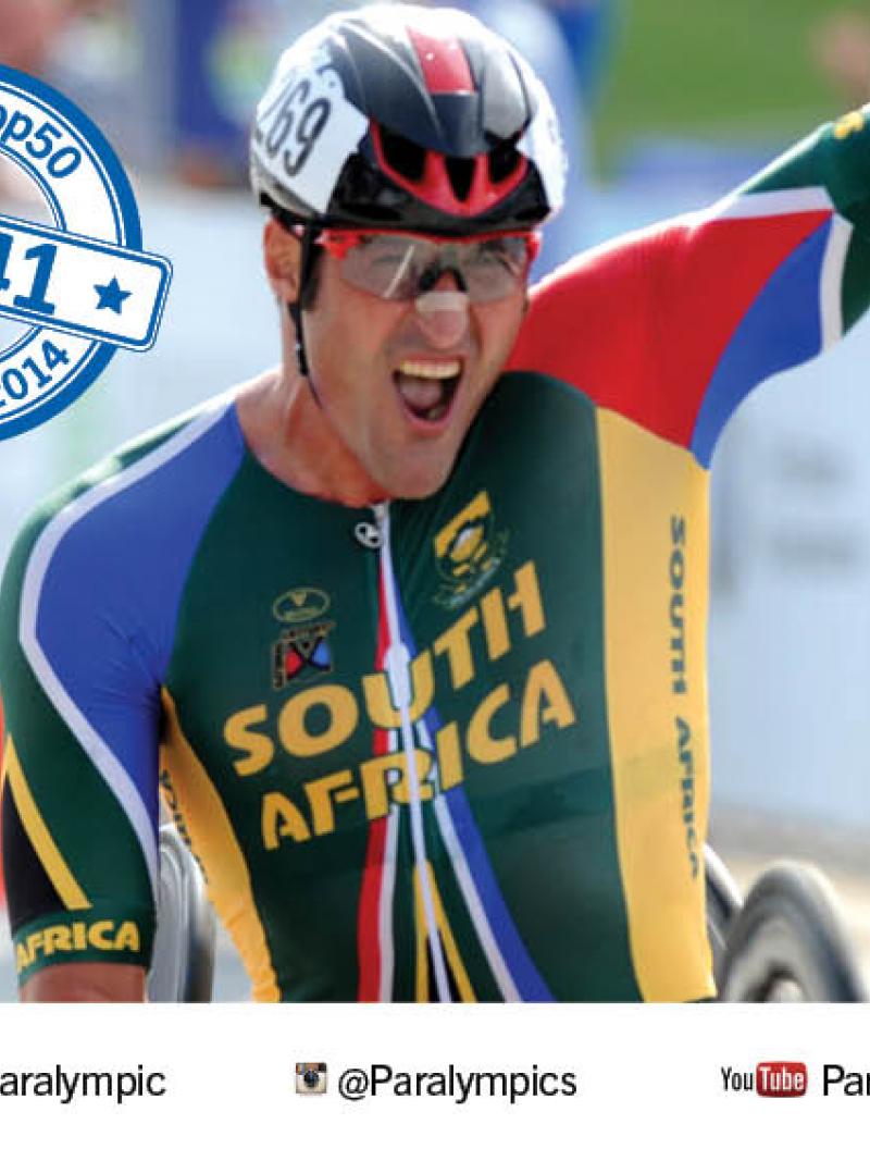 Two men in hand cycles cross the finish line one after another. The first one celebrates.