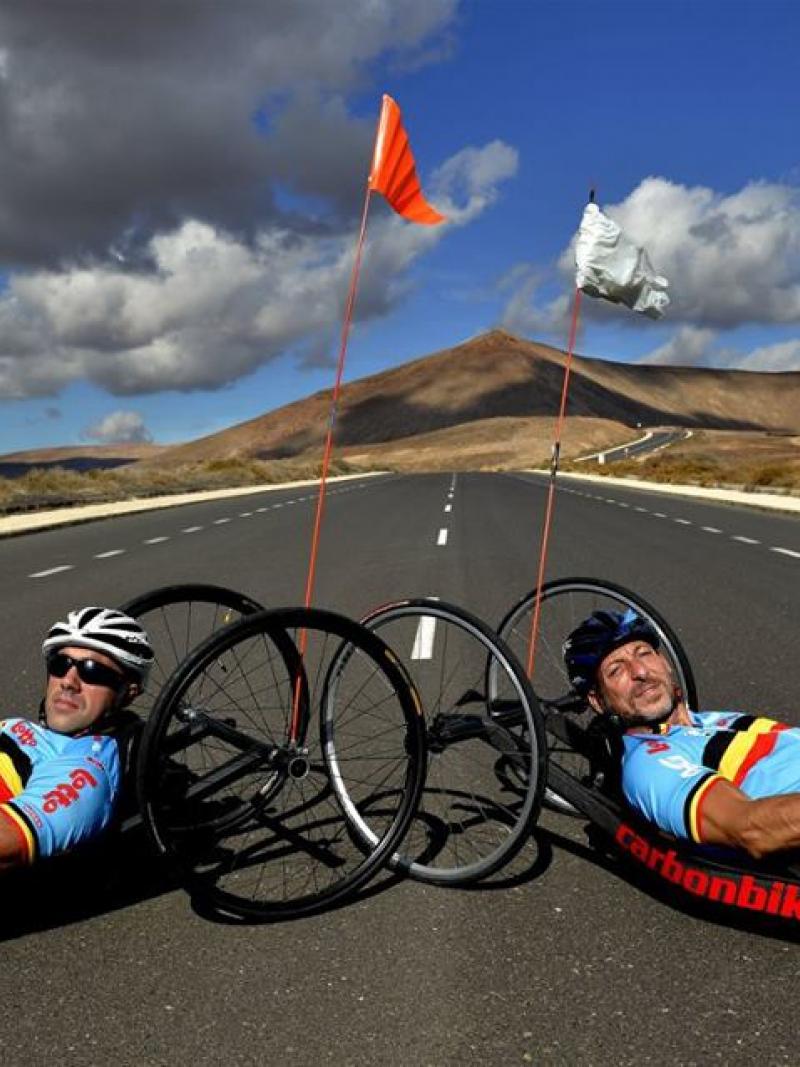 Wearing blue lycra outfits, two men lie in their handcycles on an open road with hills in the background.