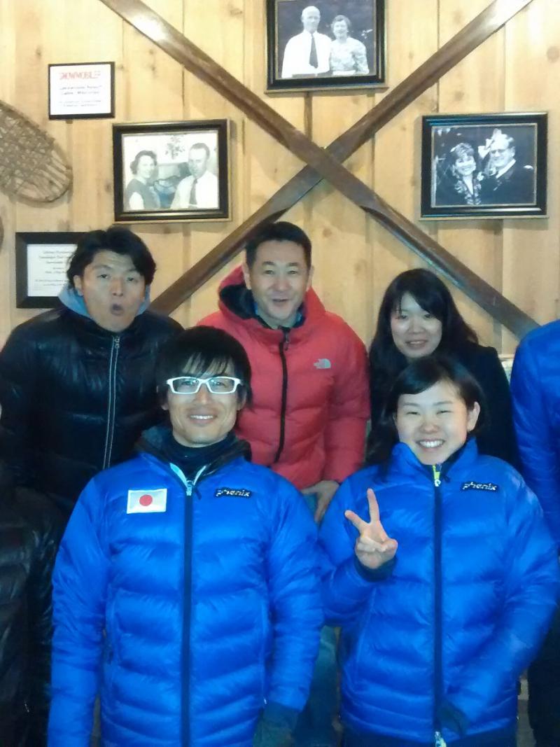 Group shot of six people in front of a wooden wall, smiling to the camera