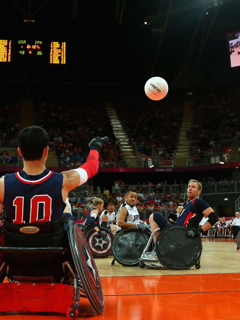Man in wheelchair throwing a ball, two other men trying to catch it