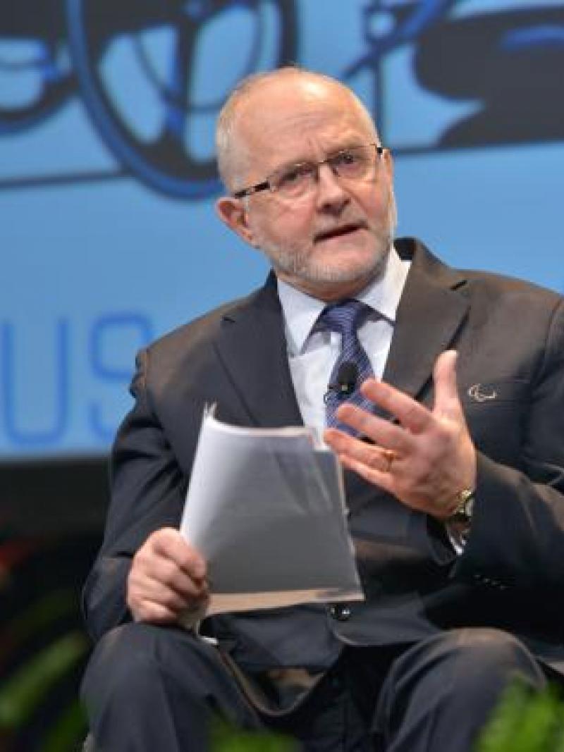 A man seated holding papers in one hand and using his other hand to express his feelings whilst giving a talk.