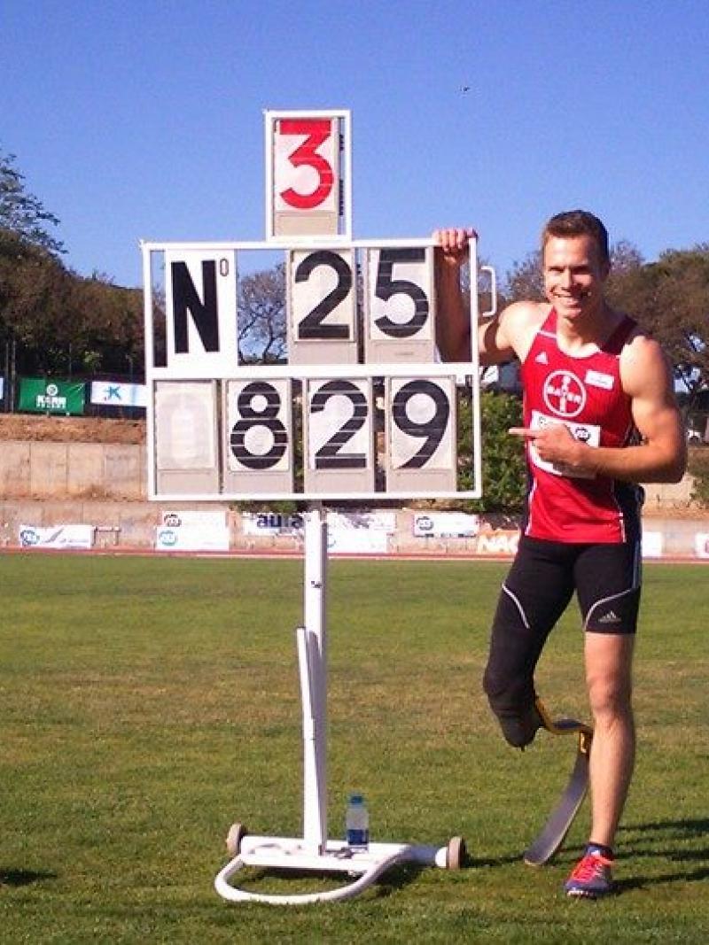 Man with prosthetic leg in a stadium, posing next to a sign that shows the number 8.29m