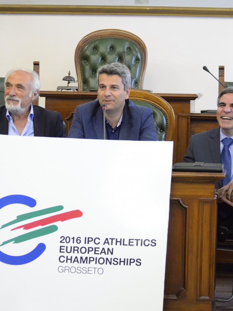 Three men behind a table present the logo for the 2016 IPC Athletics European Championships.