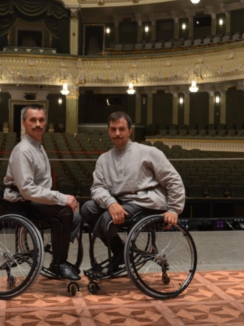 Two men in wheelchairs on a stage in front of empty seats