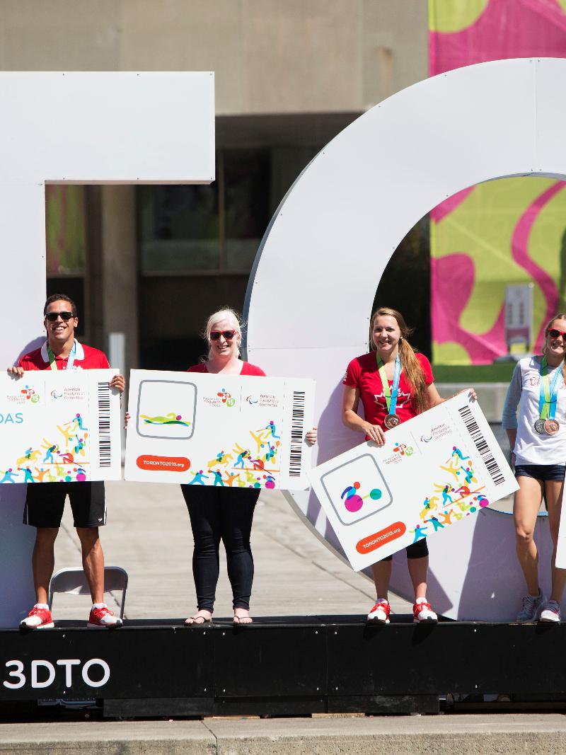 Five persons with big tickets in their hands pose in front of a big sign with the letters TO