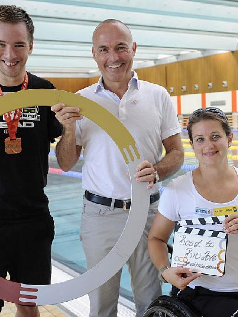 Two men and a woman in a wheelchair pose in front of a swimming pool.