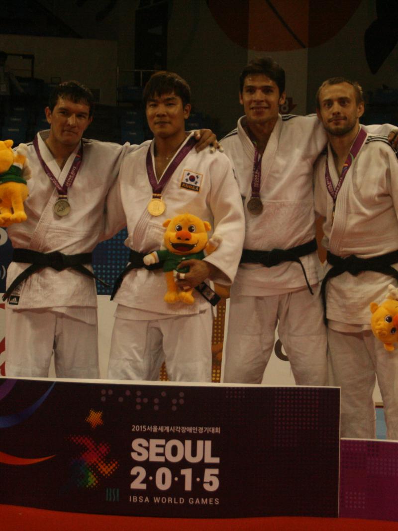Three men in white training suits stand on a podium with medals