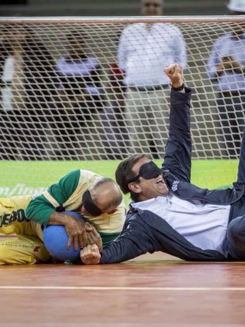 Two blindfolded men, one in a suit lying on a field of play, catching a ball