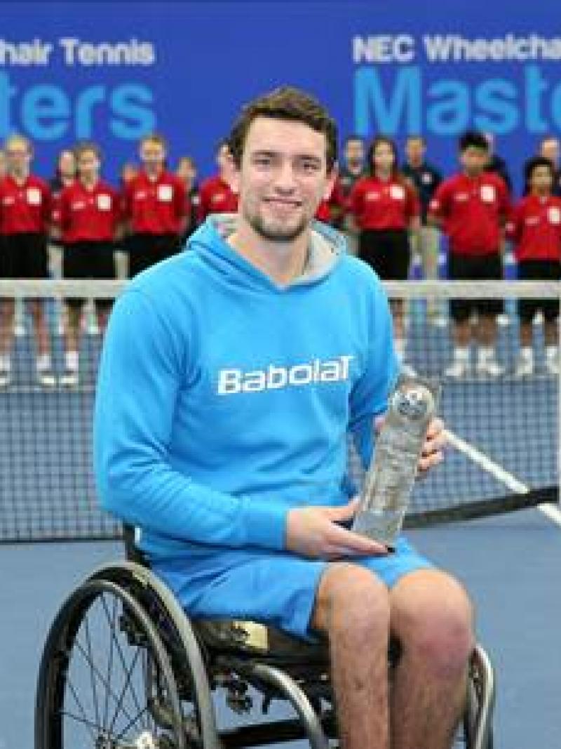 Man in wheelchair on a tennis court, showing a trophy to the camera
