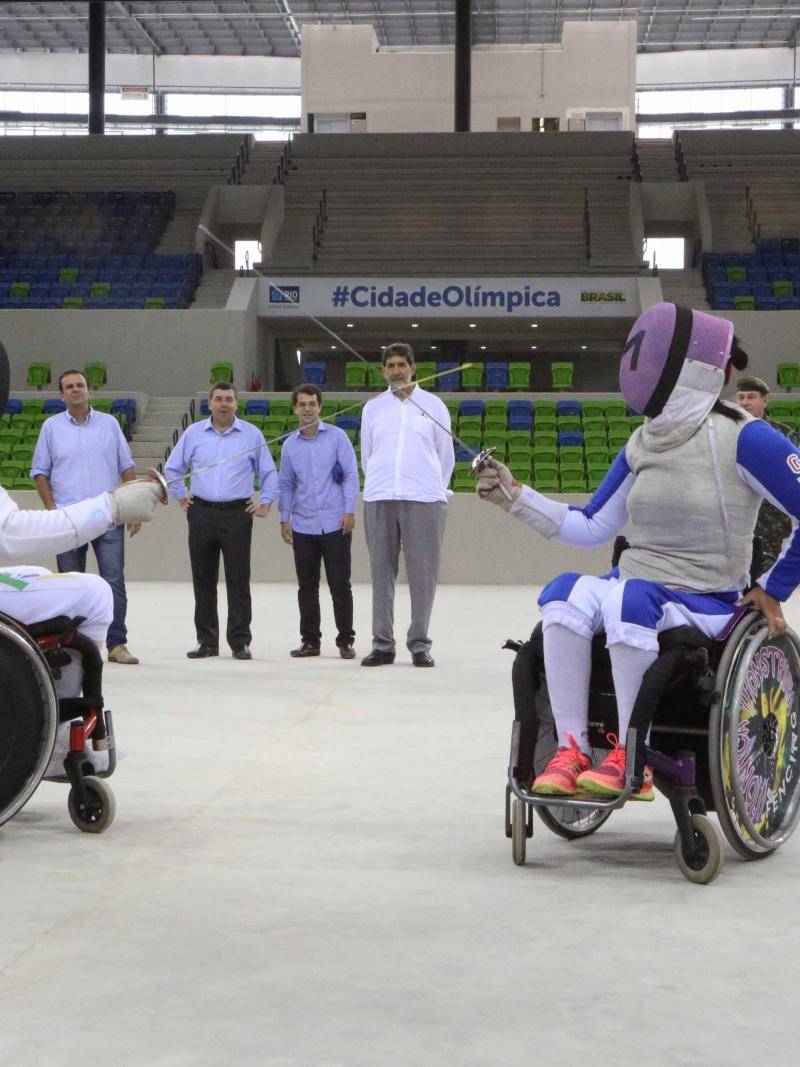 Two wheelchair fencers cross swords in empty arena, some people watching from behind