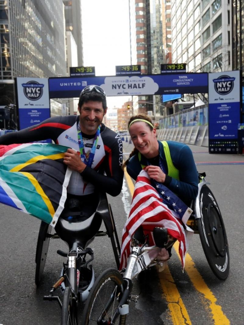 A man and a woman in racing wheelchairs, posing and showing an American and a South African flag